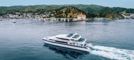 "An aerial view of Catalina Express ferry gliding through the pristine waters, with Avalon Harbor and the iconic Catalina Casino in the background surrounded by lush green hills."