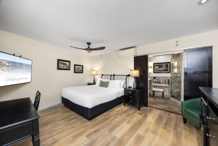 "Spacious hotel room with a large bed featuring a white quilted cover, green accent pillow, and fabric headboard with leather strap detail. The room includes wood flooring, black nightstands with matching green lamps, a black writing desk, and a wall-mounted flat-screen TV displaying 'Catalina Courtyard Suites.' A dark wood dresser with a mini-fridge and microwave sits beside a green armchair. Sliding barn-style doors open to a bathroom area with a vanity, mirror, and glass-enclosed shower. A ceiling fan with wooden blades is mounted on the white ceiling