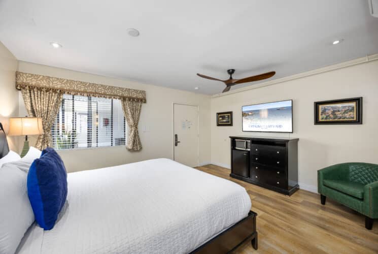 King Bed with white sheets blue pillows. Tv in the backgroudn with dresser artwork and green lounge chair