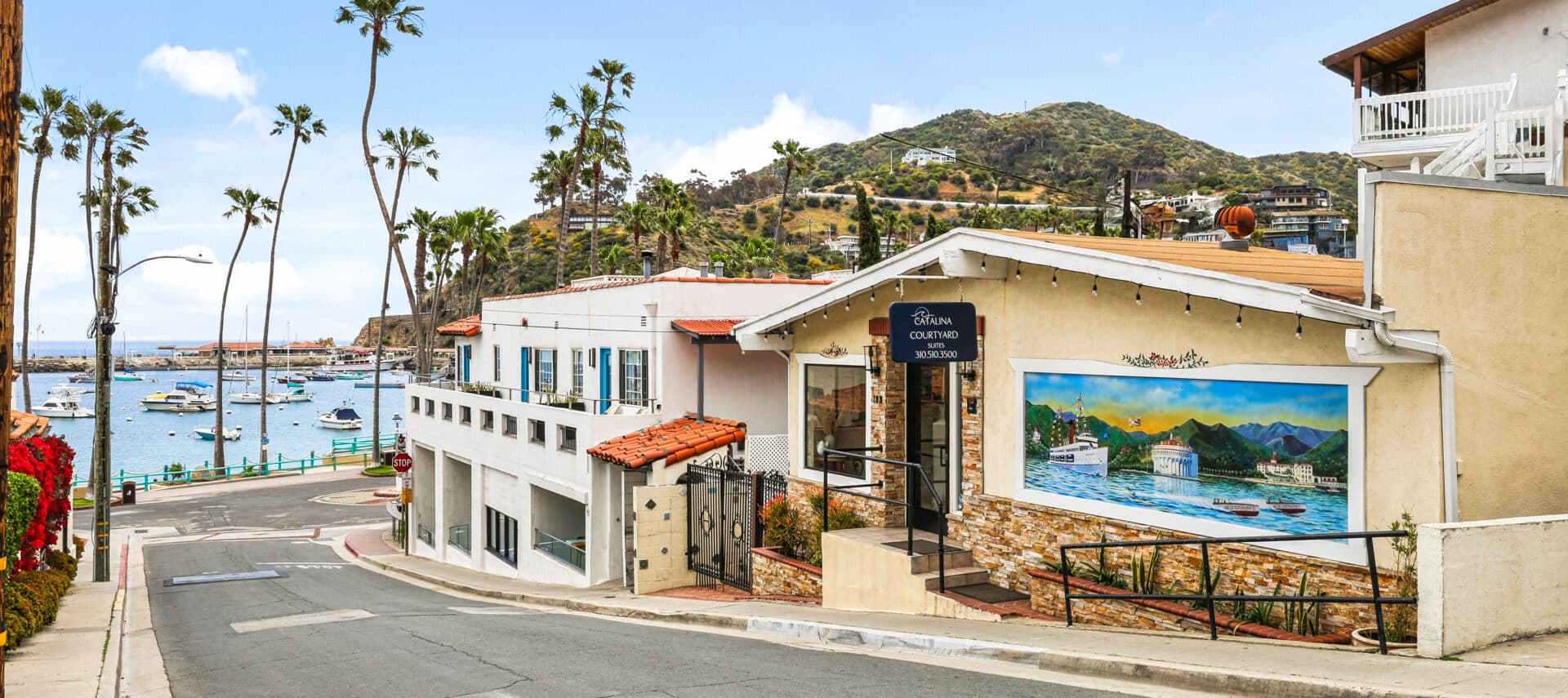 Street view of Catalina Courtyard Suites on Catalina Island, featuring a building with stone accents, a mural of the island’s harbor and Casino, and a black sign with the hotel’s name and phone number. The scene includes palm trees, hillside homes, and a marina with boats docked along the water under a clear blue sky