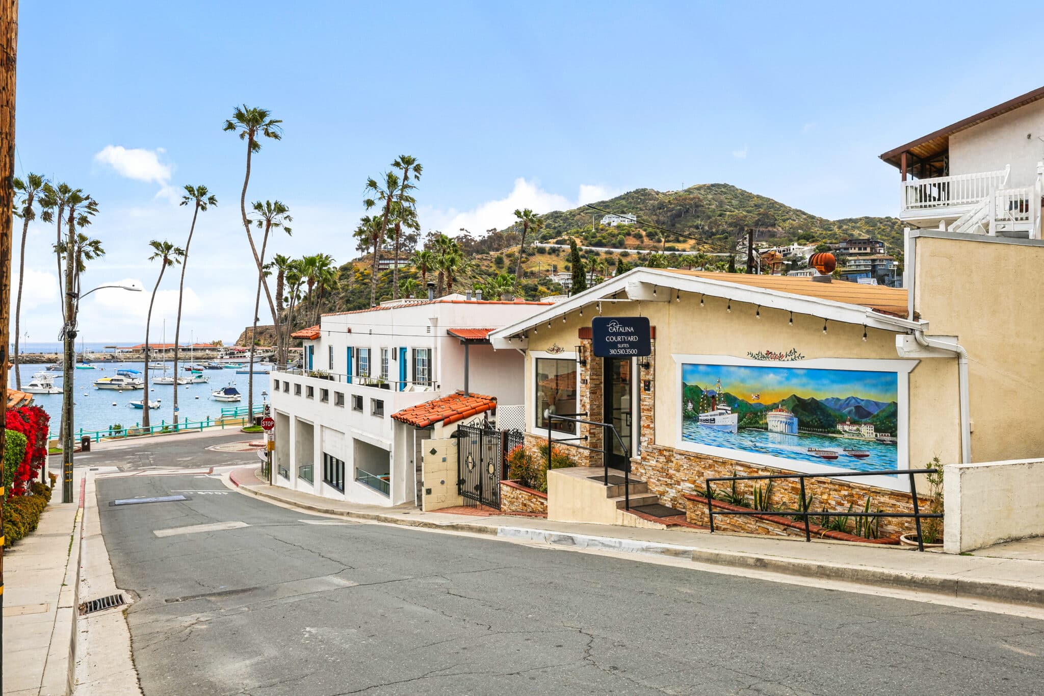 Street view of Catalina Courtyard Suites on Catalina Island, featuring a building with stone accents, a mural of the island’s harbor and Casino, and a black sign with the hotel’s name and phone number. The scene includes palm trees, hillside homes, and a marina with boats docked along the water under a clear blue sky