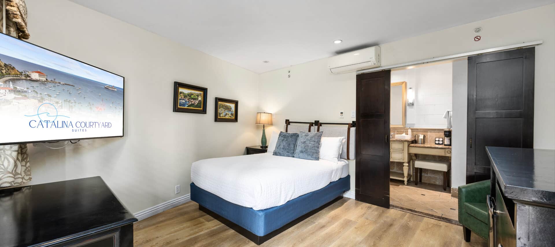 Hotel room with a neatly made bed featuring a white quilted cover and blue base, two blue accent pillows, and a headboard with leather straps. The room has wood flooring, a flat-screen TV mounted on the wall, a black desk, and framed island-themed artwork. A sliding barn-style door opens to a bathroom area with a vanity, mirror, and coffee station. A floor lamp provides warm lighting beside the bed