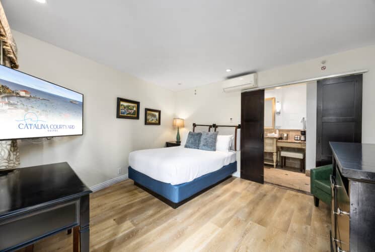 Hotel room with a neatly made bed featuring a white quilted cover and blue base, two blue accent pillows, and a headboard with leather straps. The room has wood flooring, a flat-screen TV mounted on the wall, a black desk, and framed island-themed artwork. A sliding barn-style door opens to a bathroom area with a vanity, mirror, and coffee station. A floor lamp provides warm lighting beside the bed