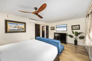 Bright hotel bedroom featuring a large bed with a white textured coverlet, a blue upholstered bench at the foot of the bed, and wood flooring. The room includes a flat-screen TV mounted above a black dresser with a microwave and mini-fridge. Nautical and island-themed artwork decorates the walls, and a large potted plant sits near patterned curtains. A modern wooden ceiling fan is mounted on the white ceiling.