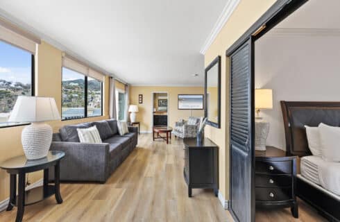 Bright and spacious Catalina Courtyard Suites hotel suite on Catalina Island featuring large windows with scenic Avalon harbor and hillside views. The suite includes a gray sofa with striped pillows, glass-top side table with a white textured lamp, wood flooring, and warm yellow walls. A separate bedroom offers a dark wood sleigh bed and nightstand behind black louvered sliding doors. Additional features include two patterned armchairs, a round coffee table, and a flat-screen TV. Ideal Catalina Island lodging near Avalon beach, harbor, and local attractions.