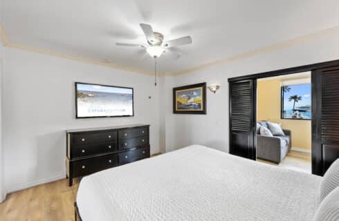 Catalina Courtyard Suites hotel room on Catalina Island featuring a bright bedroom with a large bed, white quilted bedding, black dresser, flat-screen TV, nautical artwork, and ceiling fan. Sliding doors open to a sitting area with a gray sofa and ocean views of Avalon harbor and cruise ships. Ideal Catalina Island lodging offering ocean-view accommodations near Avalon beach and local attractions.