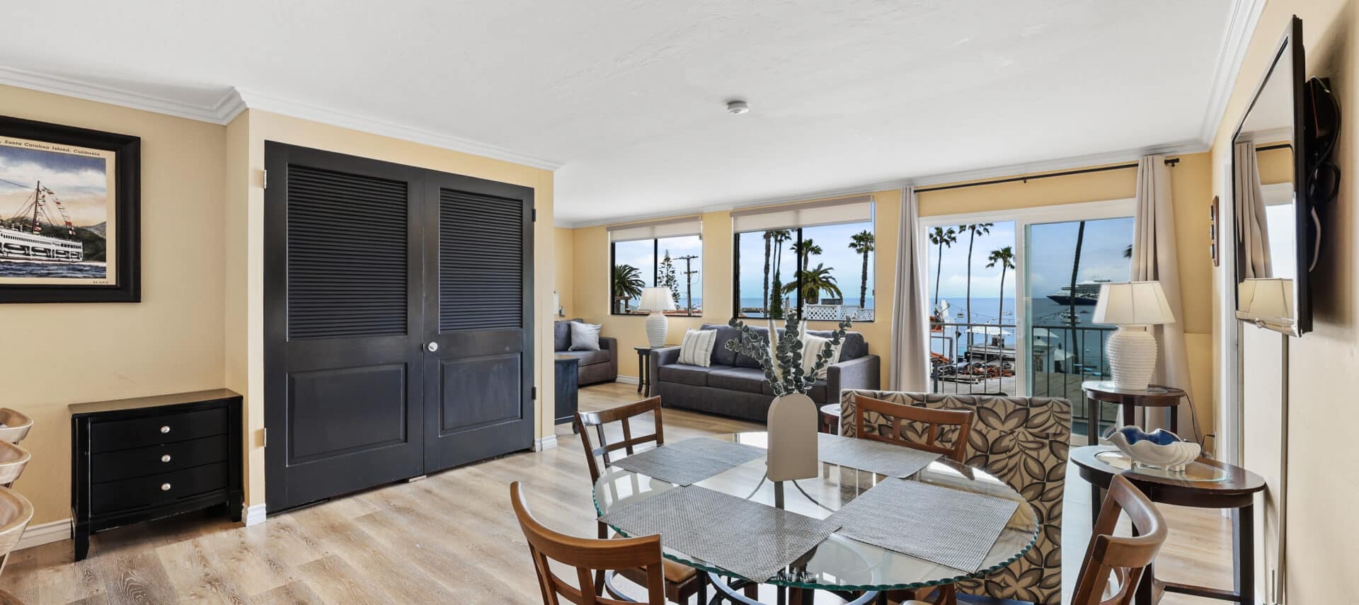 Spacious ocean-view suite at Catalina Courtyard Suites featuring a dining area with a glass table, cozy seating area with two sofas, large windows, and sliding glass doors offering stunning views of Avalon Bay and palm trees. The room is decorated in warm tones with modern coastal furnishings and natural light throughout.