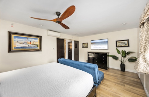Bright hotel bedroom featuring a large bed with a white textured coverlet, a blue upholstered bench at the foot of the bed, and wood flooring. The room includes a flat-screen TV mounted above a black dresser with a microwave and mini-fridge. Nautical and island-themed artwork decorates the walls, and a large potted plant sits near patterned curtains. A modern wooden ceiling fan is mounted on the white ceiling.