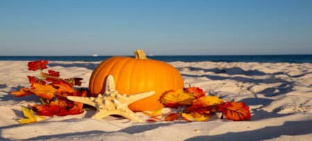 "Autumn-themed beach scene featuring a bright orange pumpkin, colorful fall leaves, and a white starfish resting on soft sand with the ocean and blue sky in the background."