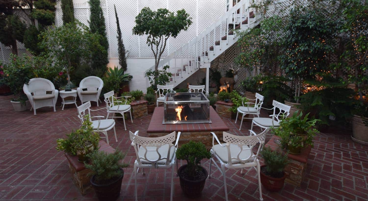 Courtyard patio with red brick, white chairs around a stone fire pit with greenery and sparkling lights around