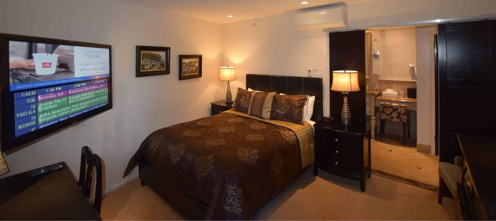 Dark wooden bed with brown and tan bedding, dark wood nightstands, and large flat-screen TV mounted on wall
