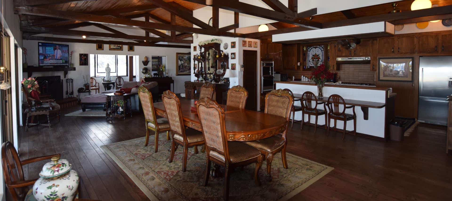 Very large living space with wood floor, ceiling, and beams including large ornate wooden dining table, kitchen area, sitting area, fireplace, and large flat-screen TV