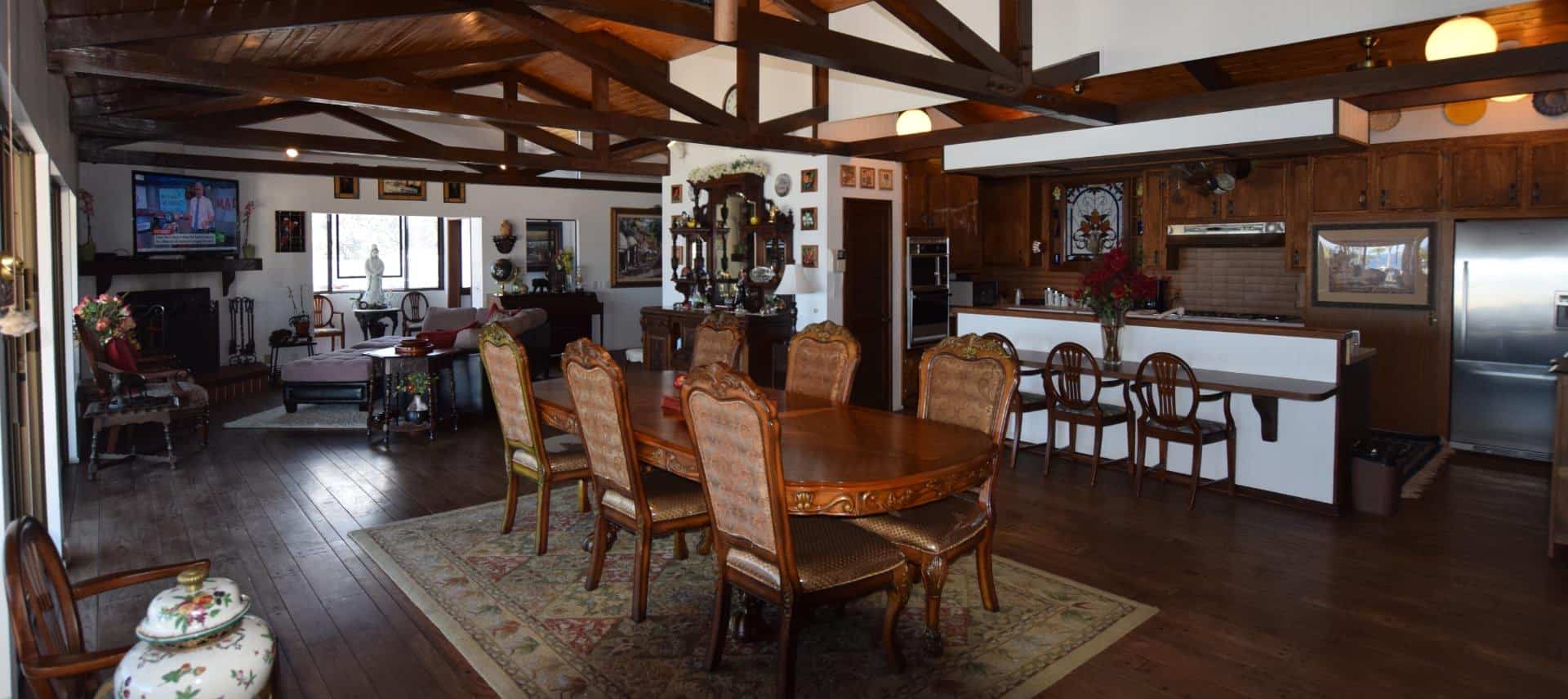 Very large living space with wood floor, ceiling, and beams including large ornate wooden dining table, kitchen area, sitting area, fireplace, and large flat-screen TV