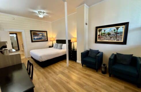 Hotel room featuring a large bed with a white quilted cover, white and green accent pillows, and a fabric headboard with leather strap details. The room has wood flooring, black nightstands with matching green lamps, and a black writing desk with a chair positioned under a window dressed with patterned gold curtains and blinds. A flat-screen TV mounted near the window displays 'Catalina Courtyard Suites,' and framed artwork decorates the walls. A wooden-blade ceiling fan is mounted on the white ceiling.