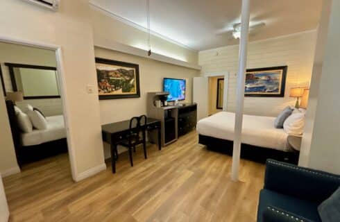 Hotel room featuring a large bed with a white quilted cover, white and green accent pillows, and a fabric headboard with leather strap details. The room has wood flooring, black nightstands with matching green lamps, and a black writing desk with a chair positioned under a window dressed with patterned gold curtains and blinds. A flat-screen TV mounted near the window displays 'Catalina Courtyard Suites,' and framed artwork decorates the walls. A wooden-blade ceiling fan is mounted on the white ceiling.